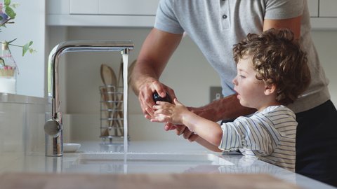Father helping young son to wash hands in kitchen sink during health pandemic - shot in slow motion