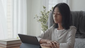 Happy Asian girl waving in greeting online tutor via internet communication on tablet while sitting in room at home. Concept online learning at home