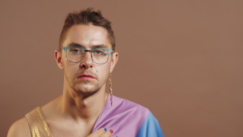 Chest up portrait shot of young stylish gay man wearing makeup, dress and rainbow flag over his shoulder holding hand on chest and looking at camera while posing in studio