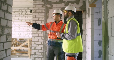 Zoom out view of contractor demonstrating construction site and talking with male engineer with tablet inside unfinished brick building