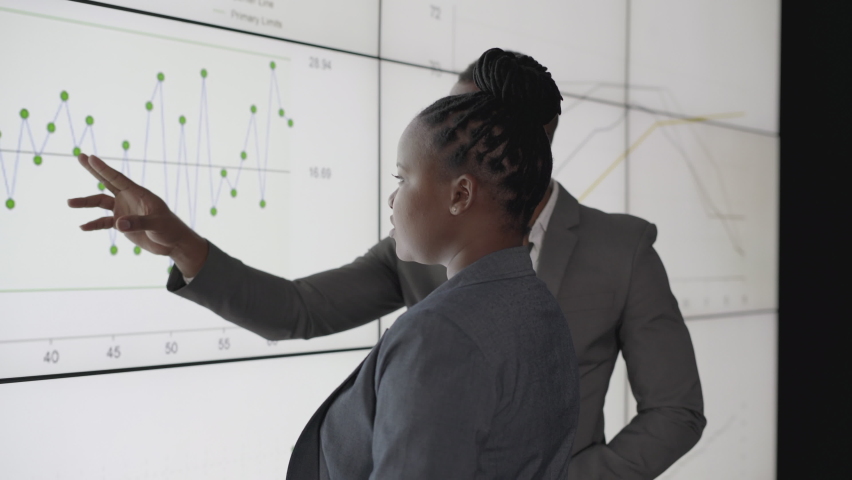 Two young African businesspeople discussing data
together on a digital multiscreen wall during a
presentation to coworkers in a dark office Royalty-Free Stock Footage #1061837056