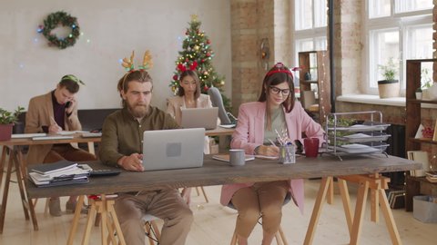 PAN of man with beard wearing Christmas antlers headband sitting down at desk in open plan office. He is turning on his laptop and looking at screen. People working in office on Christmas