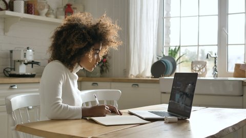 A cheerful African-American girl listens and takes notes on an online lecture from her teacher, online education at home due to quarantine. A mixed race girl with a lush hairstyle is studying at home