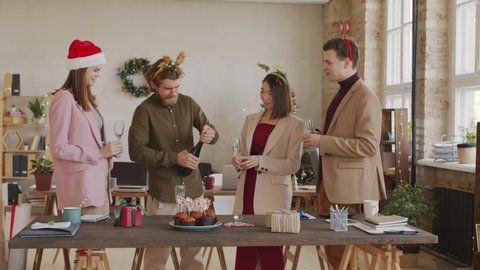 Medium shot of happy bearded man in festive headband opening bottle of champagne and pouring it into glasses of his cheerful colleagues while celebrating Christmas with them in office Vídeo Stock