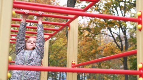 Little girl swinging on playground monkey bars hanging on arms in autumn park