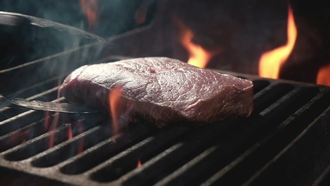Juicy raw beef steak is fried fire coals iron grill on dark background in flame of smoke. Barbecue in evening, chef turns meat on grill with tongs. cooking dinner open fire in bonfire flame.