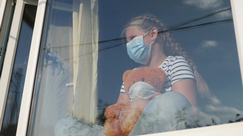 bored kid in medical mask in home quarantine coronavirus sitting by the window. child with a toy teddy bear in protective masks looking out the window. coronavirus epidemic prevention concept covid 19