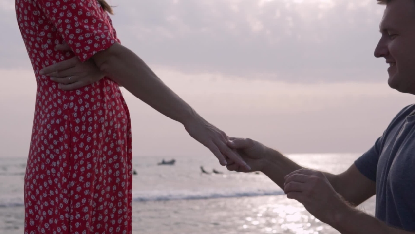 A man stands on his knee and puts a ring on his ring on a ring finger against the sea at sunset. A man proposes a hand and a heart to his woman.