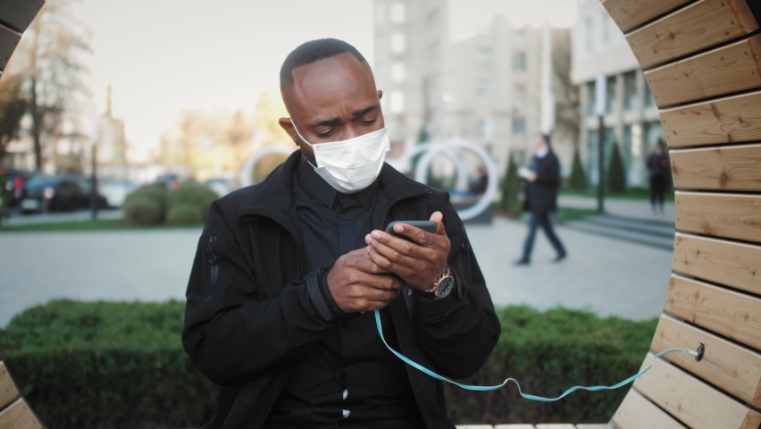 Smiling afro american millennial man hold smartphone watching social media stories video sit on bench with USB mobile device charging in the street.