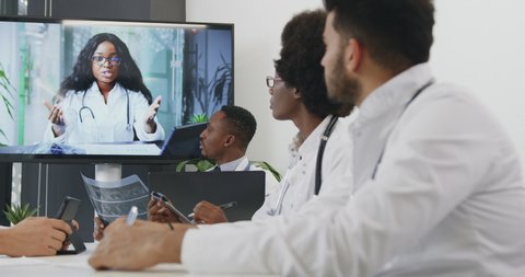 Attractive concentrated purposeful experienced diverse medical team holding online video meeting with their female colleague on interactive screen