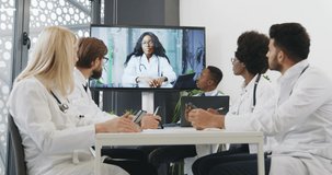 Front view of good-looking confident experienced multiracial medical team which having online video conference with female dark-skinned head doctor