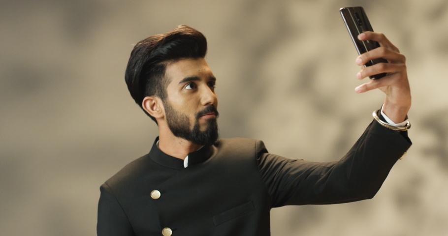 Young handsome Hindu man with beard in blac jacket smiling cheerfully and taking selfie photo on smartphone camera. Attractive happy Indian male posing to phone cam and making selfies pictures.