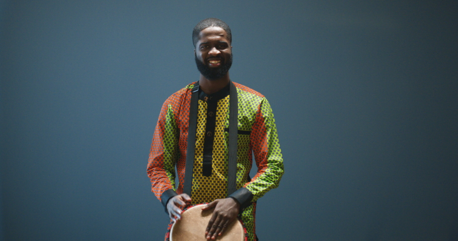 Portrait of male African American musician in glasses tapping on drum and smiling to camera. Handsome man in traditional clothes playing on bongo with happy face. Musician with musical instrument.