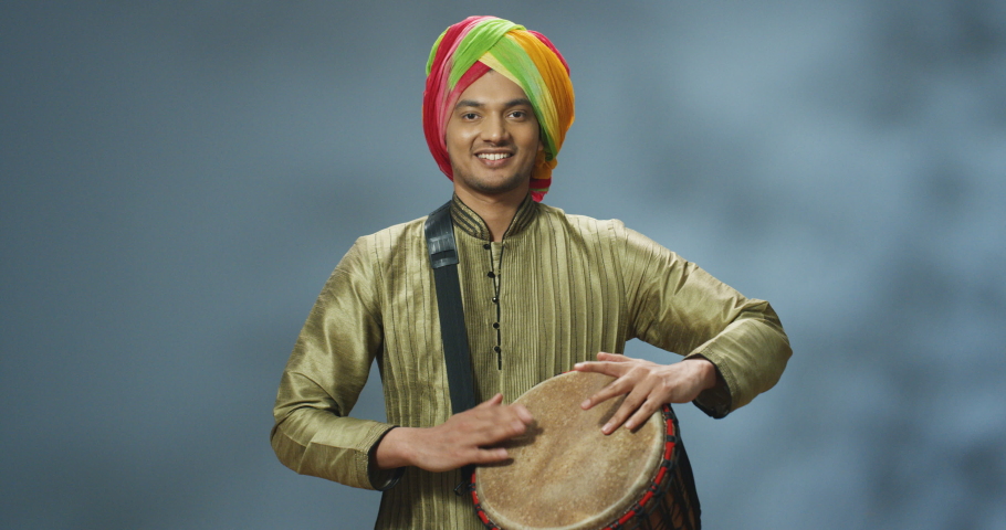 Portrait of male Indian musician in turban tapping on drum and smiling joyfully to camera. Handsome man in traditional clothes playing on bongo with happy face. Musician with musical instrument.
