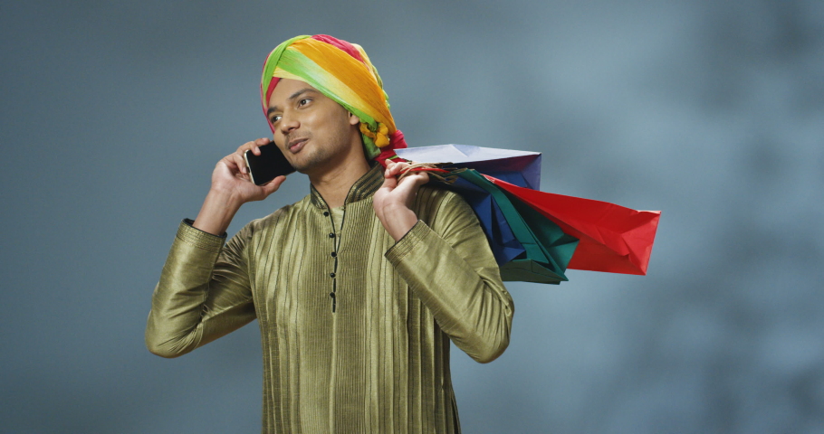 Joyful smiled young Hindu man in turban on head talking on mobile phone and holding packets over shoulder after shopping. Handsome guy speaking on cellphone with smile and bags with buyings in hand.