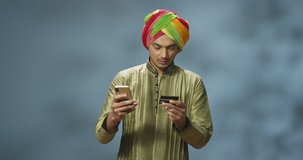 Handsome cheerful Hindu guy in turban shopping online on smartphone and laughing happily. Stylish Indian young man buying via Internet and paying with credit card. Happy male buyer buy on phone.