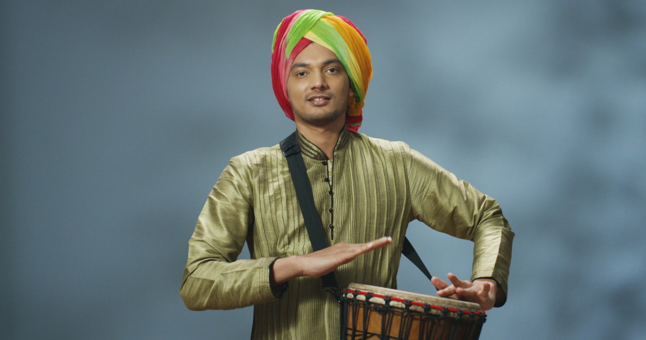 Portrait of male Indian musician in turban tapping on drum and smiling cheerfully to camera. Handsome man in traditional outfit playing on bongo with happy face. Musician with musical instrument.