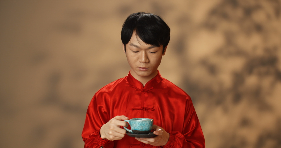 Handsome Asian young man in red traditional clothes sipping hot drink from ceramic cup and tasting it. Portrait of guy drinking tea and enjoying. Chinese tea ceremony concept.