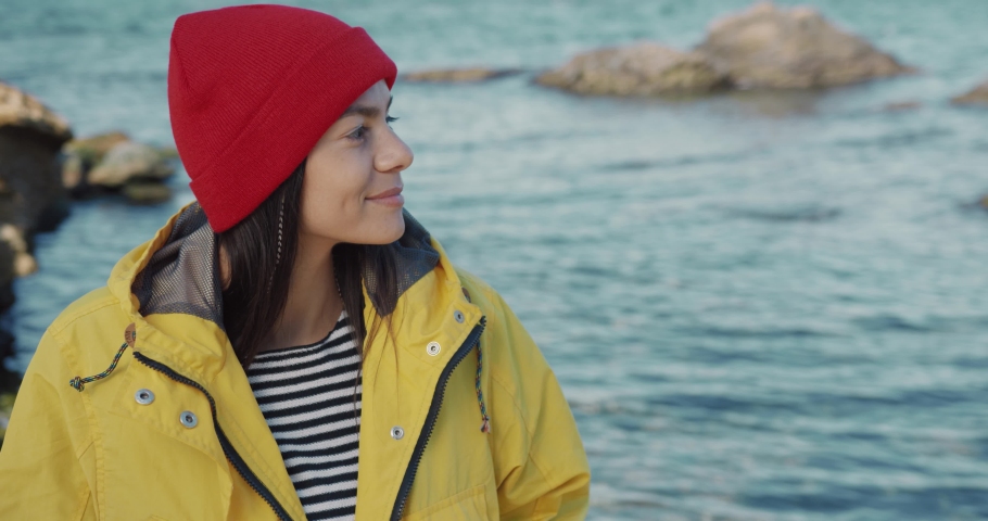 Beautiful caucasian girl inyellow jacket and red hat stands on background of sea looking at camera and smiling.