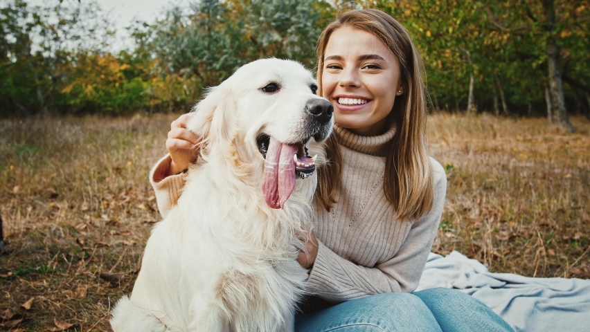 Young blonde woman in casual clothes is sitting on blanket at a glade of autumn park, smiling, hugging and stroking her funny dog labrador retriever. Happy weekend with pet outside. Close up | Shutterstock HD Video #1061863117