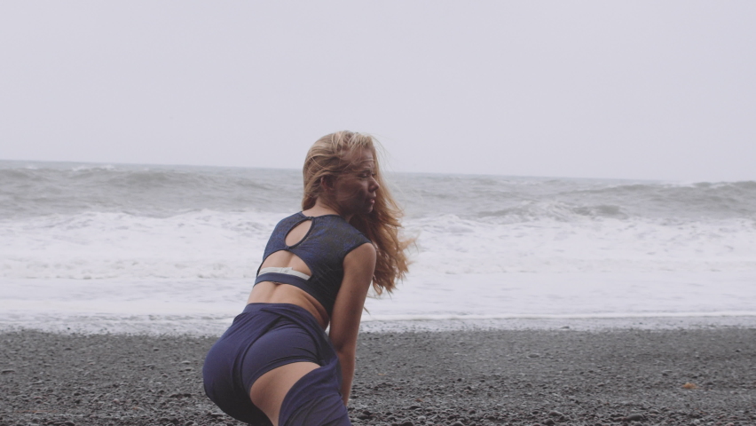 Panoramic view of the black sand and oceanscape view with a woman and her arms held high, knees bent, as she dances and enjoys the cold breeze.