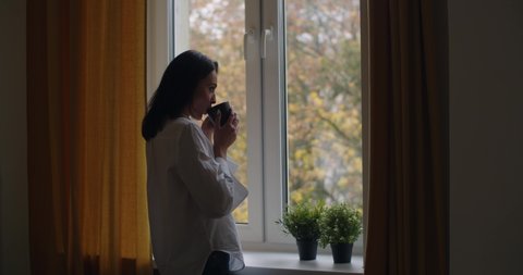 Side portrait of pensive girl standing by window with cup of tea slow motion. Young woman drinking hot coffee looking outside at autumn landscape copy space. Loneliness depression solitude concept