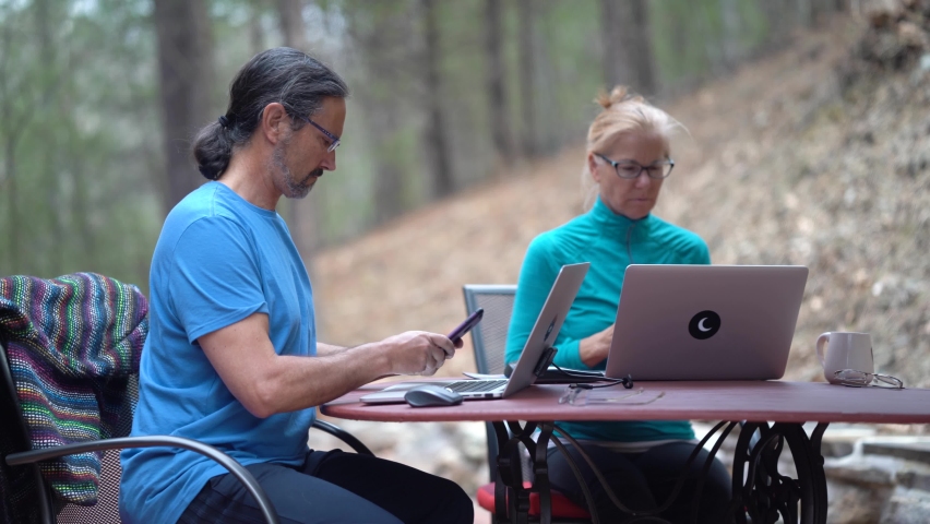 Mature man checks phone and a woman works on computer at a table outside.