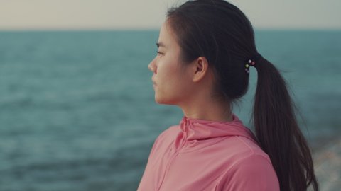A beautiful asian woman runner looks at the sea view while standing on the beach. Girl in sports clothes taking a break from exercising admiring the view of the beach sunset.