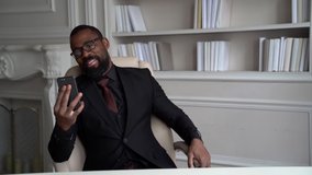 Smiling African-American bearded man in a black suit, stylish glasses. The businessman is on the desktop, sitting in a bright office. Speaks from a smartphone video.