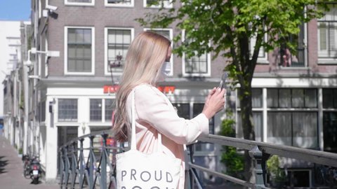 A Lovely Blonde Woman With Facemask Taking Selfies Using Her Smartphone Standing On A Canal Bridge In Red Light District, Amsterdam - Covid-19 Pandemic - Medium Shot