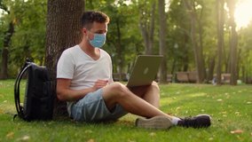 Young student in a mask in the park sits under a tree on the grass and communicates via video link