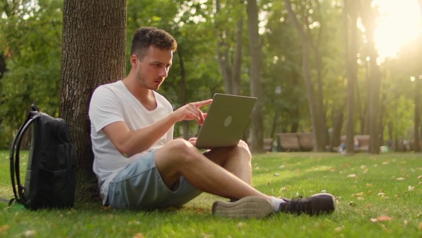Young student on the grass in the park sits under a tree and cheerfully works with a laptop Royalty-Free Stock Footage #1061887348