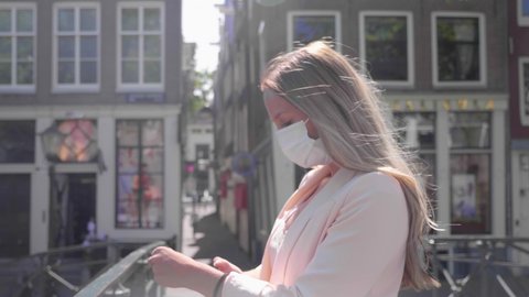 A Casual Dutch Woman Having Conversation Using Her Smartphone Wearing White Facemask On A Canal Bridge In Red Light District, Amsterdam. - Covid 19 Pandemic - Medium Shot, Slow Motion