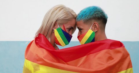 Young couple of women hugging under rainbow flag wearing colorful masks at gay pride event - Social distance and love concept - Slow Motion