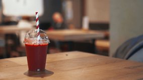 Smooth circling 4k view of a red smoothie in a plastic cup with a red and white striped straw on a wooden restaurant table with blurry people and the interior of a restaurant as background