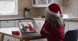 Caucasian woman spending time at home wearing santa hat, sitting in kitchen having video chat with friends on laptop screen, waving in slow motion. self isolation at christmas time during covid 19