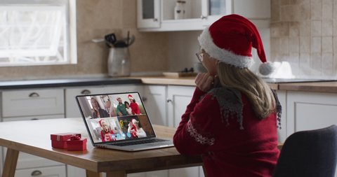 Caucasian woman spending time at home wearing santa hat, sitting in kitchen having video chat with friends and family on laptop screen, waving in slow motion. self isolation at christmas time
