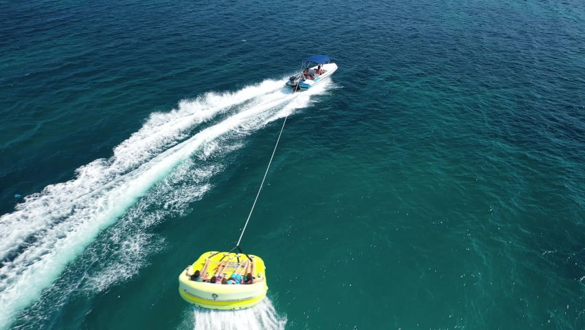 Aerial drone video of extreme inflatable towable tube cruising in high speed attached on powerboat with children on sofa ride, watersport Royalty-Free Stock Footage #1061900161