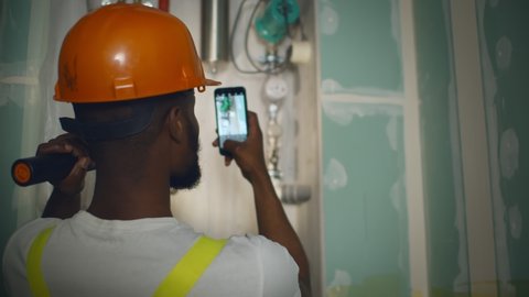 Back view of african engineer with flashlight taking photo on smartphone of broken heating system. Afro-american construction worker sending photo of boiler system to plumber