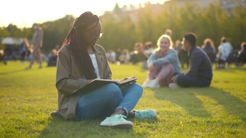 Happy african student lady reading book outdoors in park. Portrait of cheerful afro college female sitting cross-legged on campus lawn preparing for exam
