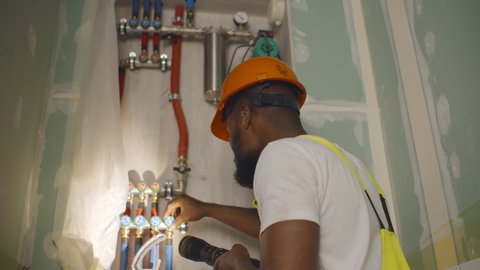 Back view of african professional fixing house central heating system. Afro engineer in uniform and hardhat setting plumbing system in new house.