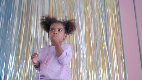 Little African American girl is eating and also a bit dancing in front of twinkling curtain with gold and silver color and she look happy to enjoy eating in her home.