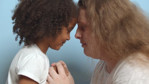 Side view of caucasian woman and mixed race daughter looking at each other isolated over blue background. Close up of adoptive mother and preschool girl cuddling