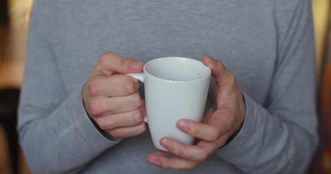 Front view of male hands holding white cup indoors slow motion close up. Faceless man drinking tea enlightened with morning natural light. Breakfast energy healthy lifestyle. Healthcare habits