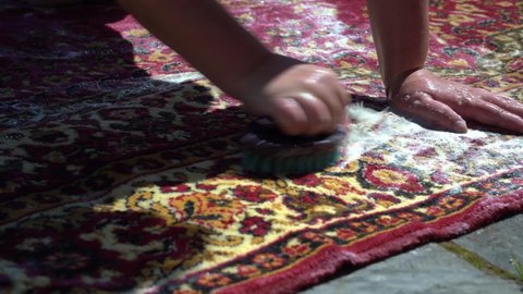 Washing carpet with foam and water by hand outside. A woman cleans a dirty carpet in the street, in the courtyard of a house on a stone floor. Close-up. Rinse with clean water.