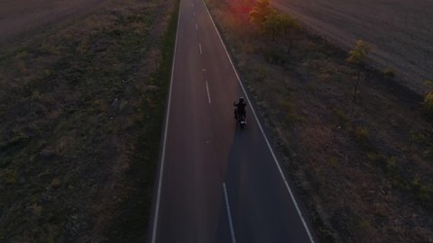 aerial shot, motorcyclist in safety helmet rides bike on road reflecting suns rays at sunset through fields after harvest
