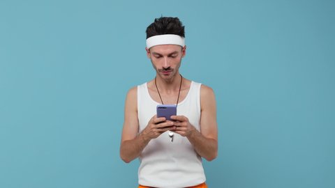 Excited young fitness man sportsman in headband shirt isolated on blue background. Workout gym sport motivation concept. Using pointing finger on mobile cell phone typing sms message showing thumb up