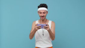 Crazy cheerful young fitness man with skinny body sportsman in headband shirt isolated on blue background. Workout gym sport motivation concept. Play game on mobile phone screaming put hand on head