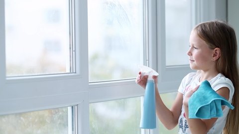 A little girl sprinkles water from a bottle on the window and wipes it off with a rag. The child helps with cleaning the house. Wash the window.