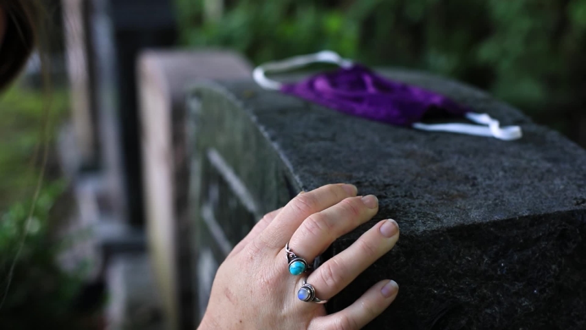 Selective focus of young woman hand touching tombstone at cemetery mourning for deceased during covid-19 pandemic outbreak with mask lying on stone Royalty-Free Stock Footage #1061918458